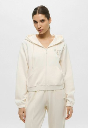 Felpa con zip di colore crema, dotata di cordino regolabile, tasche laterali e polsini a coste; presenta un logo ricamato sul petto. Abbinata a pantaloni coordinati.