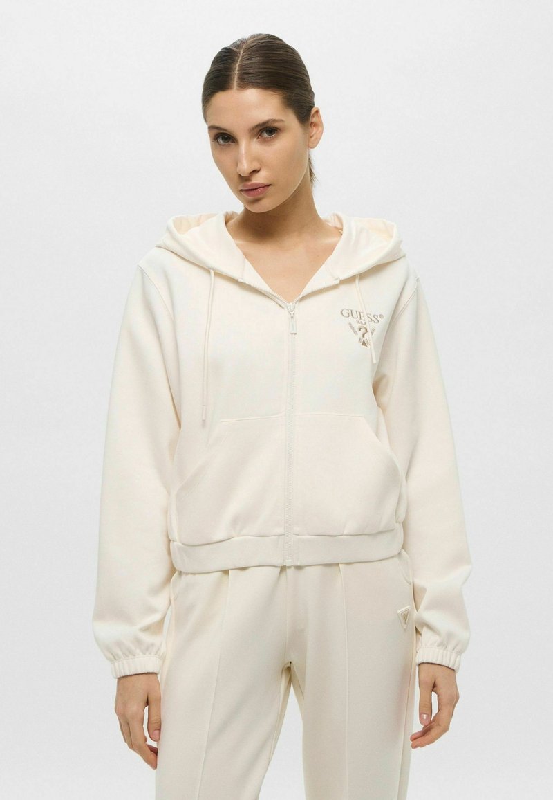 Felpa con zip di colore crema, dotata di cordino regolabile, tasche laterali e polsini a coste; presenta un logo ricamato sul petto. Abbinata a pantaloni coordinati.