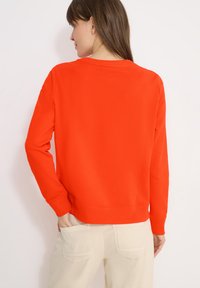Femme aux cheveux bruns portant un pull à manches longues orange vif et un pantalon beige clair, debout avec une main dans une poche arrière.