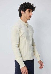 Homme aux cheveux bouclés et foncés portant un polo à manches longues crème à côtes et un pantalon sombre, se tenant devant un fond gris clair.