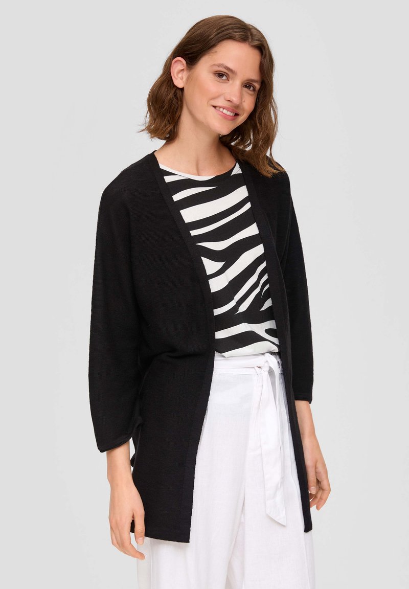 Schwarze offene Strickjacke mit langen Ärmeln, getragen über einem weißen und schwarzen gestreiften Oberteil, kombiniert mit weißen Hosen mit einer Krawattenbund.