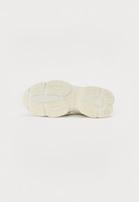 Suela de zapatilla blanca con una superficie texturizada, que presenta patrones ovalados para el agarre y un detalle circular central. Material de goma, diseño ligero.