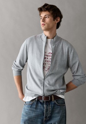 Jonge man draagt een lichtgrijze trui met rits over een wit bedrukt T-shirt en blauwe spijkerbroek, staand met zijn handen achter zijn rug.