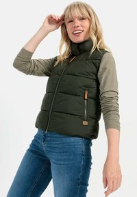 Grüne Steppweste mit hohem Kragen, Frontreißverschluss, zwei Seitentaschen und braunen Lederakzenten. Getragen über einem langärmligen grauen Hemd und blauen Jeans.