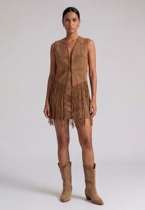 Donna che indossa un gilet senza maniche in camoscio marrone e una minigonna con frange coordinata con stivali da cowboy color cuoio, in piedi su uno sfondo semplice.