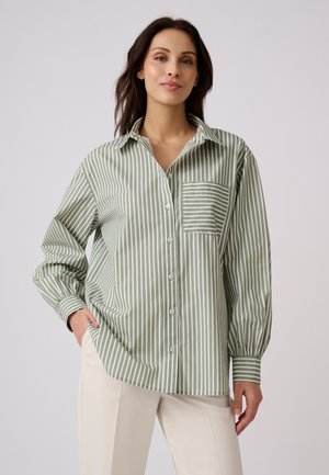 Donna che indossa una camicia larga a righe verticali verdi e bianche con bottoni, con una mano in tasca e pantaloni di colore neutro su sfondo semplice.