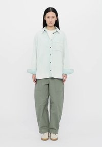 Cămașă albastră deschis cu mânecă lungă și buzunar frontal, asortată cu pantaloni cargo largi de culoare verde măsliniu. Modelul poartă pantofi crem, stând în fața unui fundal alb.