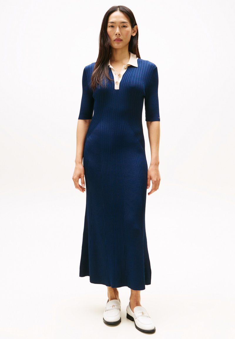 Granatowa sukienka dzianinowa z prążkowanym wzorem, o dopasowanym kroju, z krótkimi rękawami i kremowym kołnierzykiem z guzikami. Uzupełniona białymi butami wsuwanymi.
