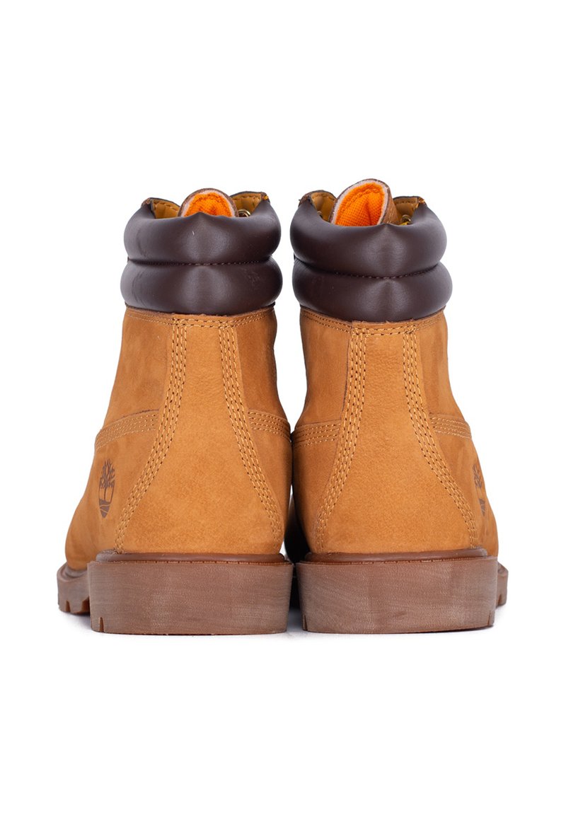 Un paio di stivali alla caviglia in pelle color tan con colletto imbottito marrone scuro, rivestimento interno arancione e suole in gomma spessa, mostrati da dietro.