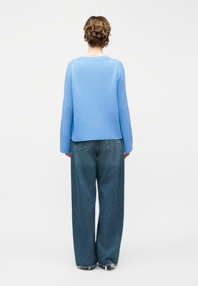 Άτομο με κοντά, σγουρά μαλλιά που φοράει γαλάζιο πουλόβερ και άνετο σκούρο μπλε τζιν, στέκεται με την πλάτη γυρισμένη σε απλό φόντο.