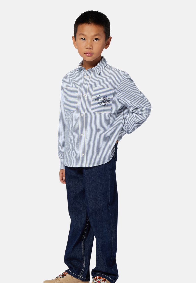 Gestreept shirt met lange mouwen in blauw en wit, met een voorzak met geborduurde tekst, gecombineerd met donkerdenim broek en lichte schoenen.