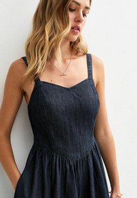 Denim jurk in donkerblauw met een vrouwelijke pasvorm en een uitlopende rok, voorzien van brede schouderbanden en een hartvormige halslijn. Vloeiende textuur en minimale afwerking.