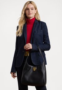 Musta nahkalaukku, jossa on kaareva muoto, vetoketjukiinnitys ja kultaiset yksityiskohdat. Kantaa olalla denimmanttelin ja punaisen poolopaidan kanssa.