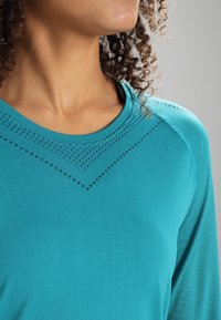 Person mit lockigen Haaren, der ein türkisfarbenes Langarmshirt mit einem dezenten Zickzackmuster auf der Brust und an den Schultern trägt.
