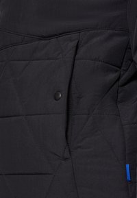 Närbild av svart quiltat jacka med diagonala sömmar, som visar en tryckknappsficka och en liten blå tygetikett på sid sömmen.