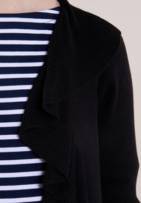 Svart ribbad cardigan med en volangkrage som hänger över en marinblå och vit randig topp, vilket visar upp ett texturerat tyg och lagerdesign.