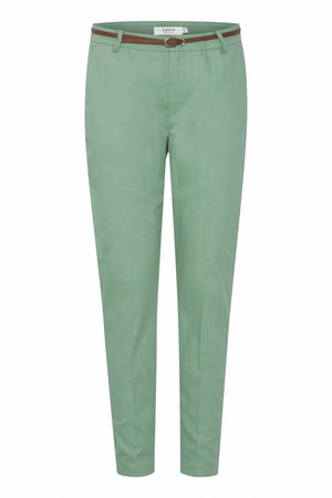Personne portant un pantalon vert sur mesure avec des poches arrière et une fine ceinture marron, associé à une chemise blanche à manches longues.