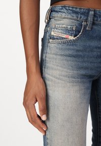 Jeans i denim med en bleknad blå färg, med en främre ficka som har en etikett med texten "Diesel Industry," och en närbild av en hand som vilar på tyget.