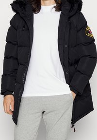Chaqueta negra tipo puffer con capucha forrada de pelo, cierre de cremallera y automáticos, bolsillos frontales y un parche con el logo en la manga izquierda.