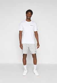 White short-sleeve t-shirt with a black logo on chest, paired with gray shorts featuring a logo on hem. Model wears white shoes and socks.
