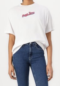 Bílá bavlněná tričko s kulatým výstřihem a krátkými rukávy, s barevným logem "Guess Jeans" přes hrudník. Nosit s modrými džíny.