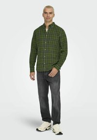 Zielona kraciasta flanelowa koszula z czarnymi liniami, z guzikami z przodu i jedną kieszenią na piersi, w połączeniu z ciemnymi dżinsami i jasnymi sneakersami.