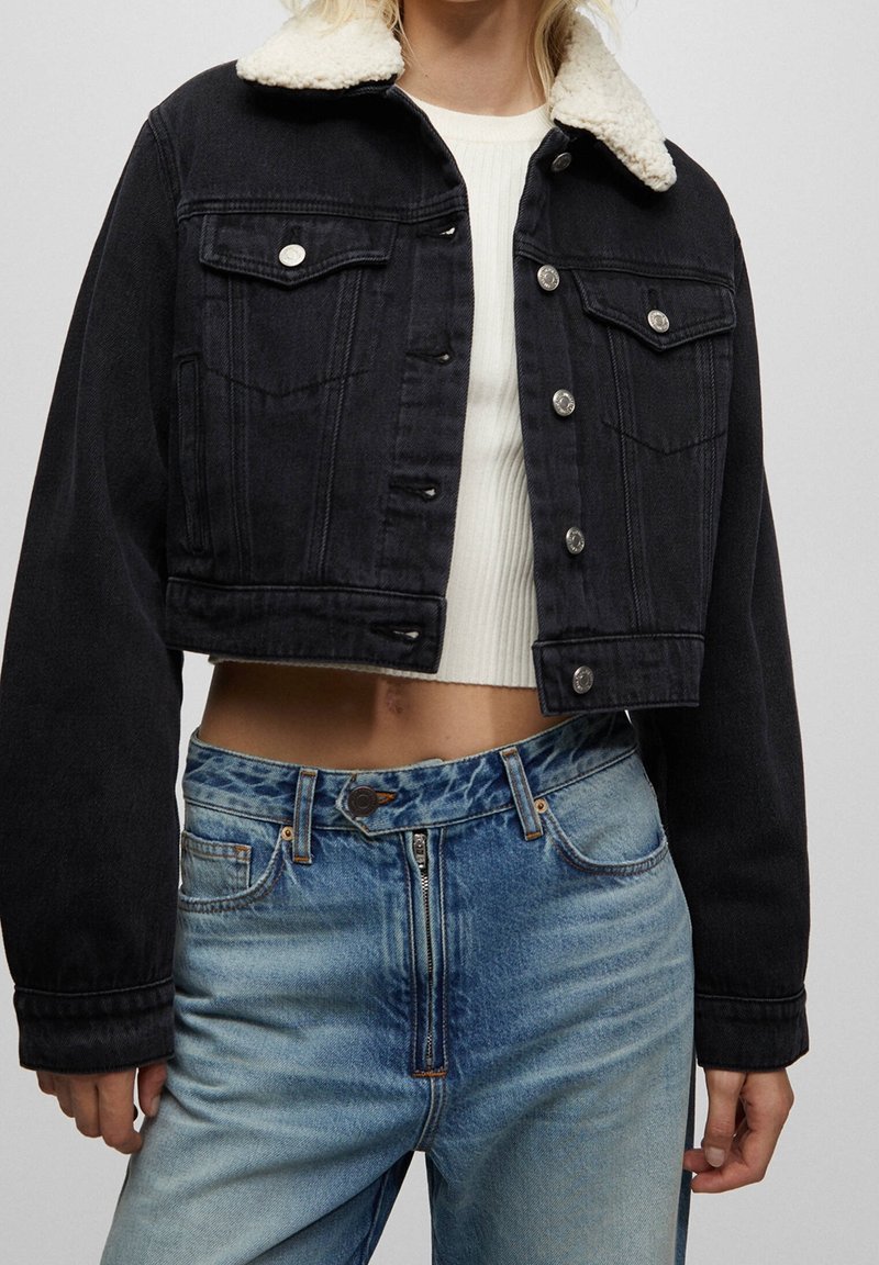 Veste en jean noire courte avec col en fausse fourrure crème, boutons argentés et deux poches poitrine, associée à un jean bleu taille haute.