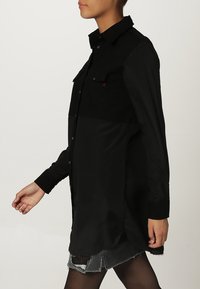 Camicia nera con bottoni, dalla finitura lucida e con dettaglio della tasca, abbinata a una gonna in denim nera strappata. Presenta maniche lunghe e una vestibilità rilassata.