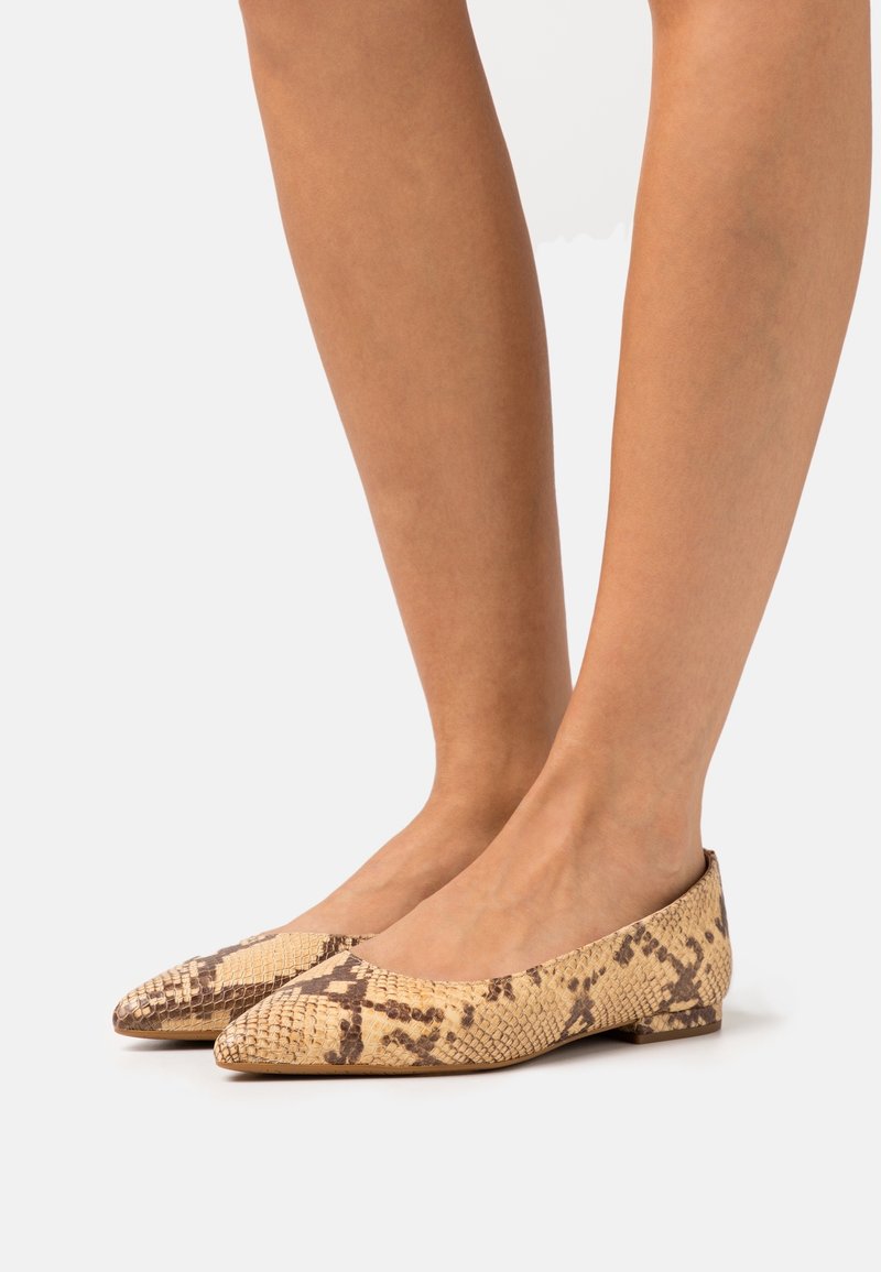 Personne portant des chaussures plates à bout pointu avec un motif peau de serpent beige et marron sur fond clair.
