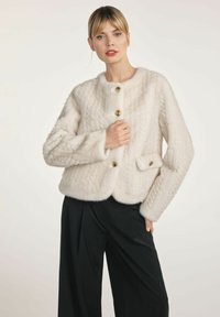 Biała futrzana kurtka z teksturowanym wzorem, okrągłym dekoltem, czterema złotymi guzikami oraz boczną kieszenią z guzikiem. W połączeniu z czarnymi spodniami.