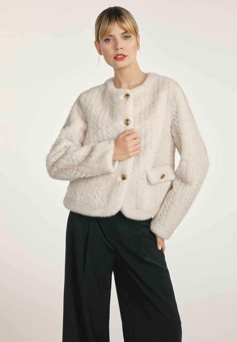 Biała futrzana kurtka z teksturowanym wzorem, okrągłym dekoltem, czterema złotymi guzikami oraz boczną kieszenią z guzikiem. W połączeniu z czarnymi spodniami.