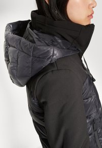 Veste noire avec une capuche matelassée et brillante. Le corps est en tissu lisse et mat. Elle possède un cordon de serrage au niveau du cou et un design ajusté.