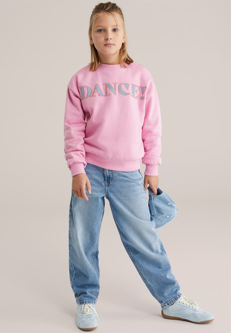 Dekle stoji sproščeno, oblečena v roza pulover z napisom "DANCE!", svetlo modre kavbojke, bele superge, v desni roki drži modro kapo.