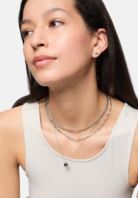 I gioielli in argento presentano catene sovrapposte, un sottile choker e un pendente con finitura opaca, indossati con una leggera maglietta a coste.