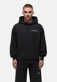 Felpa nera con cappuccio e tasca a marsupio, polsini a coste e logo "GOODFORNOTHING" in bianco. Stilisticamente abbinata a pantaloni jogger neri.