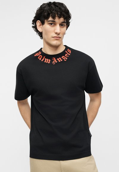 Fiatal férfi göndör sötét hajjal, fekete pólót visel, melynek nyakánál piros "Palm Angels" felirat van, kezeit a háta mögött összekulcsolva áll.