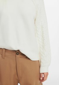 Maglione bianco a maglia con un motivo a treccia testurizzato sulle maniche, abbinato a pantaloni marroni sartoriali con dettaglio dei bottoni.
