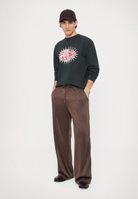 Hanorac verde închis cu un design grafic roșu, asortat cu pantaloni maro cu picioare largi. Modelul poartă o șapcă maro și pantofi negri.