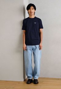 Ναυτικό μπλε βαμβακερό μπλουζάκι, ανοιχτό μπλε τζιν κανονικής γραμμής και σκούρα καφέ παπούτσια, μοντέλο στέκεται μπροστά σε έναν ουδέτερο τοίχο.