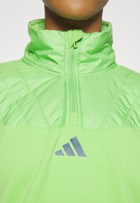 Zielona kurtka zapinana na zamek z wysokim kołnierzem, pikowaną górną częścią i gładką dolną częścią. Posiada ciemne logo i teksturowane akcenty.