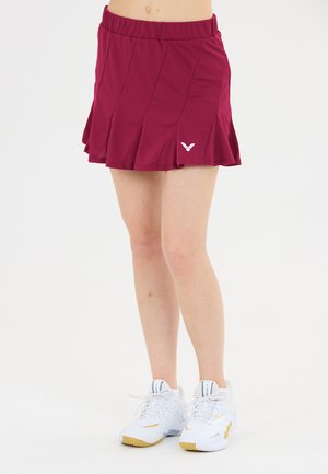 Underkrop af en person iført et plisseret burgunderrødt skørt og hvide atletikskøjter med guld accenter på en hvid baggrund.