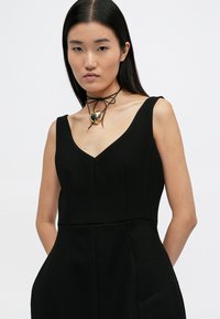 Μαύρη αμάνικη φορεσιά με V-λαιμόκοψη, διαθέτοντας εφαρμοστή κορσέ και πλαϊνές τσέπες. Διακοσμείται με δερμάτινο κολιέ και χρυσό κρεμαστό κόσμημα σε σχήμα καρδιάς.