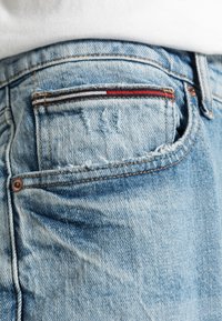 Světle modré džíny z denimu s opotřebovanou texturou, obsahující zadní kapsu s červenými a černými pruhovanými akcenty a zesíleným šitím.