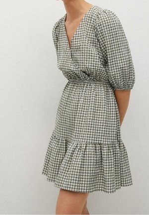 Femme portant une robe portefeuille vichy gris et blanc avec une taille froncée et des manches trois-quarts bouffantes sur un fond uni.