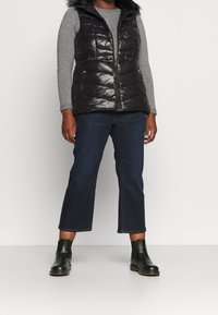 Gepolsterte schwarze Steppweste mit Kunstfellkapuze, Reißverschluss und zwei seitlichen Taschen, getragen über einem grauen Pullover und kombiniert mit dunklen, schmalen Jeans und schwarzen Stiefeln.