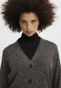 Cardigan a maglia marrone con scollatura a V, chiusura con bottoni e texture morbida, indossato sopra un dolcevita nero. Design semplice con tonalità neutre.