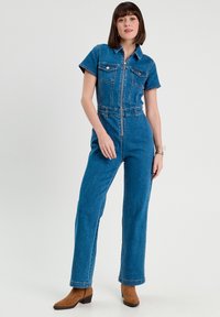 Jumpsuit in denim con maniche corte, zip frontale, tasche sul petto e vita arricciata. Presenta un colore azzurro chiaro e un design a gamba dritta.
