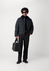 Sort pufferjakke med en tekstureret krave, båret over en sort turtleneck. Sorte bukser og polerede sko, mens der holdes en sort lædertaske.