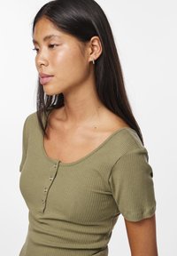 Cămașă ribbed de culoare verde măslin, cu mâneci scurte, guler în formă de cerc și cinci nasturi metalici pe față. Textură netedă și elastică.