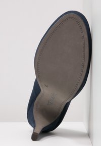Zapato de ante azul marino con puntera puntiaguda y suela de goma gris, con una etiqueta de tamaño visible "36" y el logo de la marca en relieve en la suela.